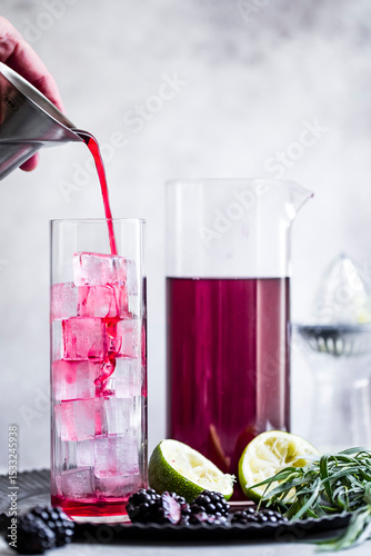 A vibrant red drink is being poured into a tall glass filled with ice cubes, surrounded by fresh blackberries, lime halves, and herbs.