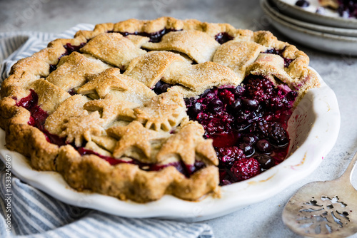 A freshly baked berry pie with a decorative crust, featuring blackberries and blueberries, sits in a ceramic dish with a slice removed.