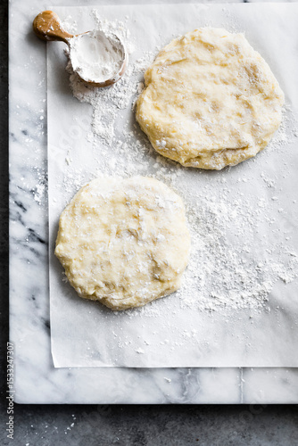 Two portions of dough on a floured parchment paper with a wooden scoop of flour on a marble surface.