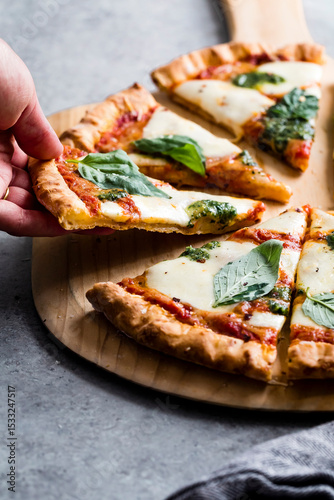 A person reaches for a slice of Margherita pizza topped with fresh basil leaves on a wooden pizza board.