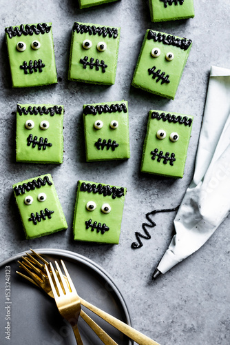 Green Frankenstein-themed cookies with black icing details are arranged on a gray surface next to a piping bag and gold forks on a plate.