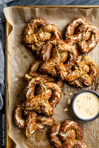 A batch of freshly baked pretzels topped with coarse salt is displayed on a parchment-lined tray, accompanied by a small bowl of creamy dipping sauce.