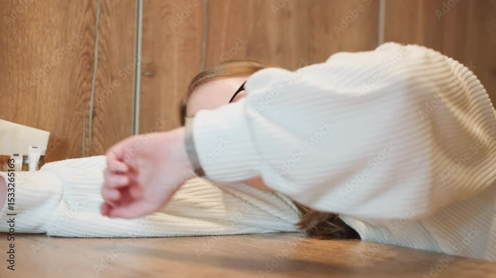 student in white sweater rests head on wooden table while arm stretches forward to check time, capturing quiet reflective moment with minimal background, napkin holder, and subtle indoor lighting