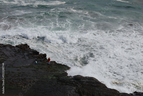 Fisherman in orange jacket fishing from a rocky outcrop, braving the large, powerful waves of the ocean at Muriwai, Auckland, New Zealand