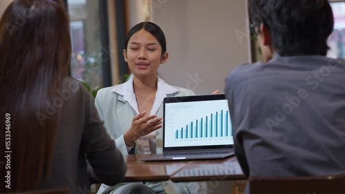 Young Asian businesswoman presenting a sales report to colleagues during a team meeting in a modern office. Confident female leader engaging in professional communication, explaining data and strategy