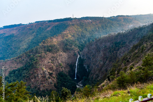 Tall, majestic Sweet waterfall of Meghalaya, cascades down a steep, rocky gorge, surrounded by lush green and autumnal vegetation.