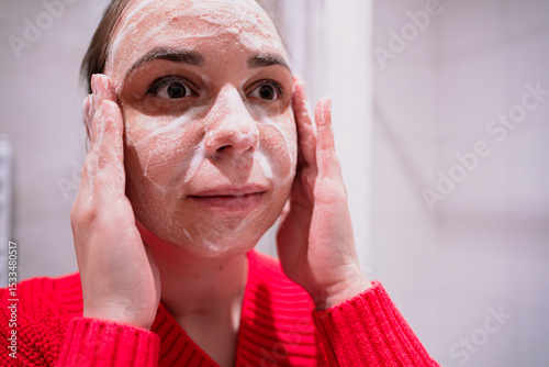 A woman is applying a cleansing product to her face while looking in the mirror She is focused on addressing her problematic facial skin and demonstrates her skin care regimen
