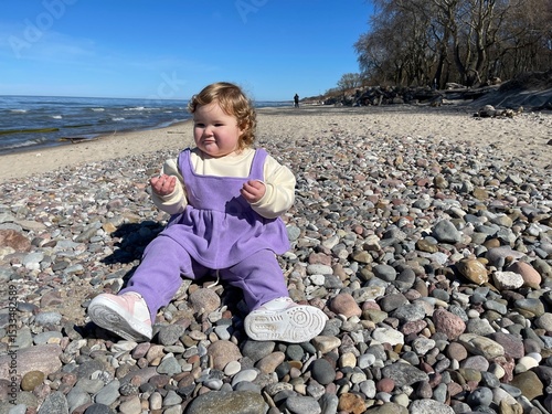 A young child sits on the beach surrounded by colorful pebbles curiously picking them up while enjoying a sunny spring day The calm water and clear sky enhance the joyful atmosphere
