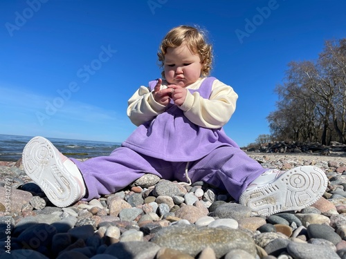 A young child sits on the beach surrounded by colorful pebbles curiously picking them up while enjoying a sunny spring day The calm water and clear sky enhance the joyful atmosphere