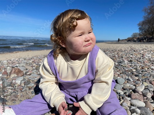A young child sits on the beach surrounded by colorful pebbles curiously picking them up while enjoying a sunny spring day The calm water and clear sky enhance the joyful atmosphere