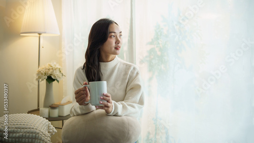 Young woman sitting peacefully at home with a cup of coffee, looking out the window.