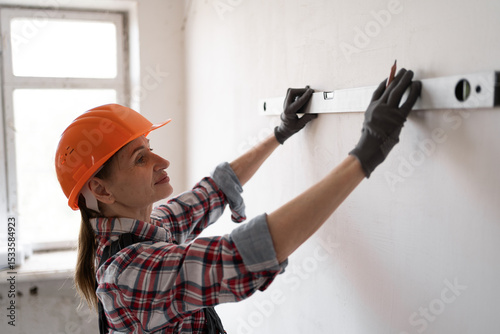 Close-up of woman builder using spirit level to measure wall and making pencil marks. Concept of renovation, construction, and skilled trade profession.