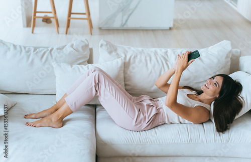 A woman lounges on a white sofa, joyfully engaging with her smartphone, in a bright and airy living space decorated in a modern, minimalist style.