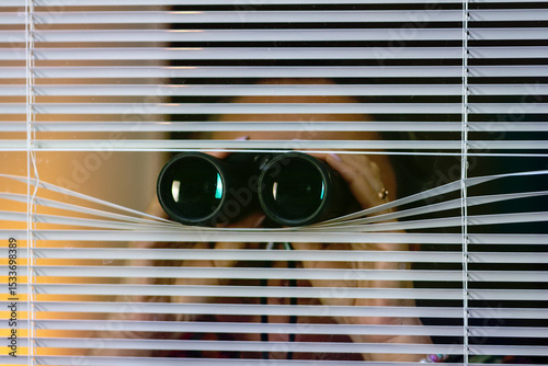 Photos Woman peeking through blinds while holding binoculars