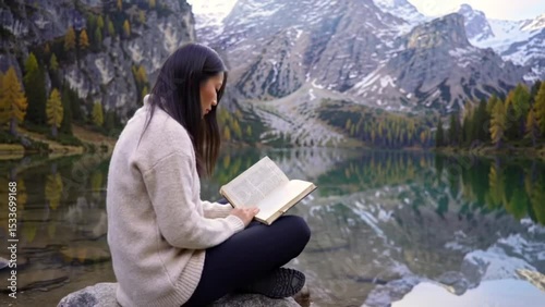 Young woman sitting on rock reading book peacefully near mountain lake with autumn trees and clear water reflection in serene landscape. Education concept
