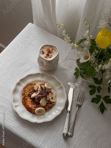 Cozy homemade breakfast - French toast with banana and chocolate, coffee, bouquet of flowers on the table in the living room by the window