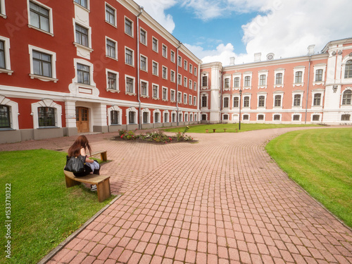 Girl on a bench in huge court yard of Jelgava palace with well looked after grass lawn and neat footpaths, blue cloudy sky. Popular city landmark and tourist attraction. Warm sunny day. Latvia
