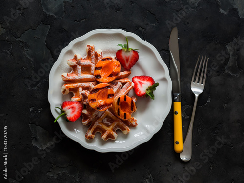 Traditional belgian waffles with caramelized apricots and strawberries - a delicious dessert on a dark background, top view