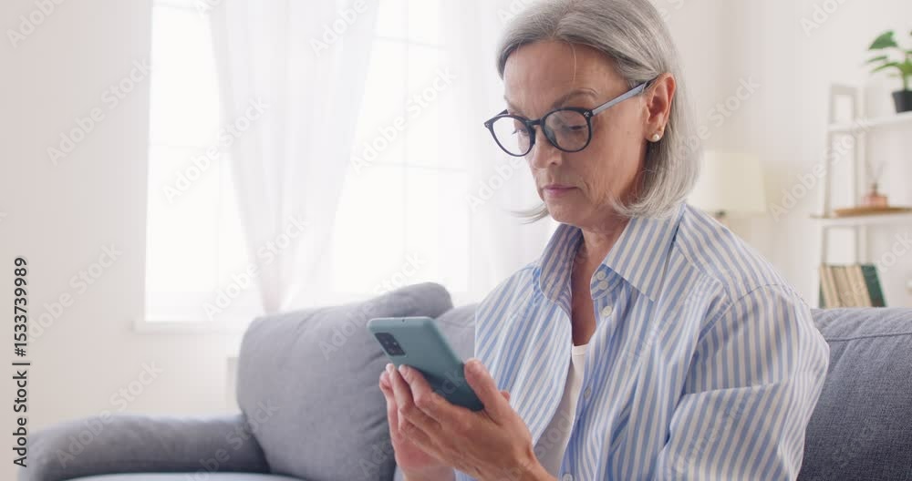 Senior woman pensioner sitting on sofa in living room at home and using smartphone browsing internet for social media. Active elderly gray-haired person using mobile phone in retirement. 4k video. Senior woman pensioner sitting on sofa in living room at home and using smartphone browsing internet for social media. Active elderly gray-haired person using mobile phone in retirement. 4k video.