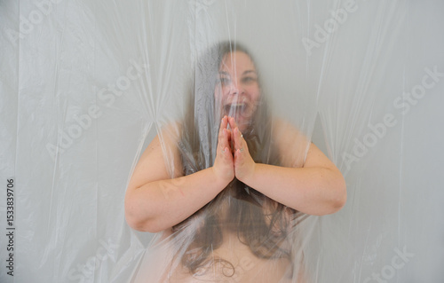 plus-sized woman is captured behind a translucent plastic sheet, her hands clasped in a fervent gesture, her wide eyes and expressive smile symbolizing unrestrained greed and desire