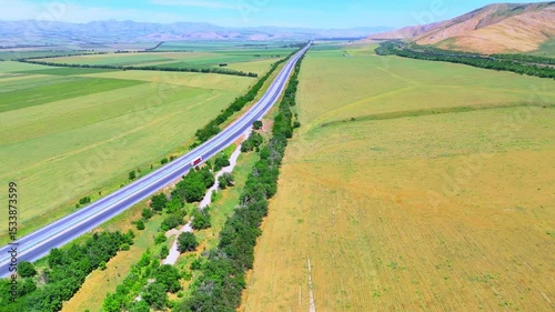 Straight highway cuts through green fields and hills. Aerial view highlights the vastness and order of the rural landscape.