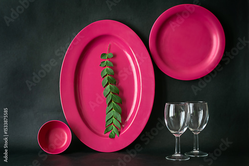 Image of a pink dinner set against dark background shot in window light. Plate is decorated with green curry laf. Set contains a serving plate, a bowl and a set of wine glasses.