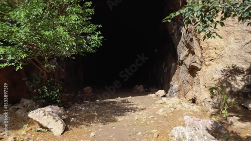 Entrance to a dark cave among rocks, surrounded by greenery and trees. The contrast of light and shadow creates a mood of mystery and untouched nature.