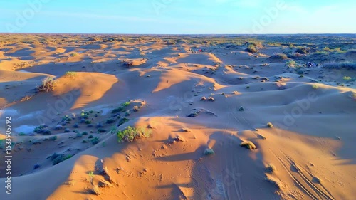 Sand dunes under a blue sky with clouds, captured by drone at sunrise. Light and shadow highlight the texture and depth of the desert landscape.
