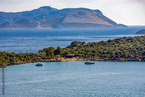 Fototapeta Naklejka Na Ścianę i Meble -  Telephoto view of Kaş peninsula: sandy beach on the narrow isthmus with sunbathers, framed by rocky cliffs and lush vegetation. Greek island rises beyond turquoise waters. Turkey, May.
