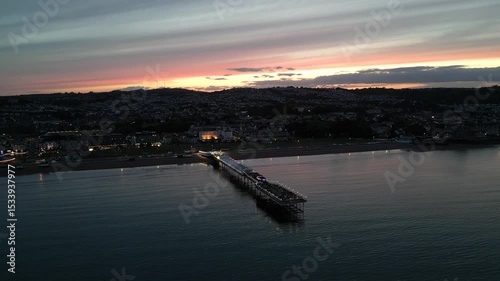 Paignton, Torbay, South Devon, England: DRONE VIEWS: Paignton Pier illuminated by a sunset. Paignton is a popular UK holiday resort and destination with many tourist attractions (Clip 1 of 3).