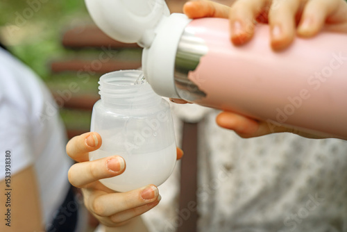 Pouring warm water from a thermos into a baby bottle to prepare infant formula during a park outing