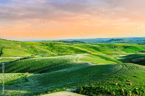 green agricultural spring or summer landscape of grassland hills near small village with young grass and amazing cloudy sky