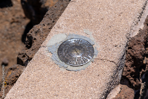 Geodetic survey marker embedded in concrete, surrounded by dark rocks, indicating a precise location.