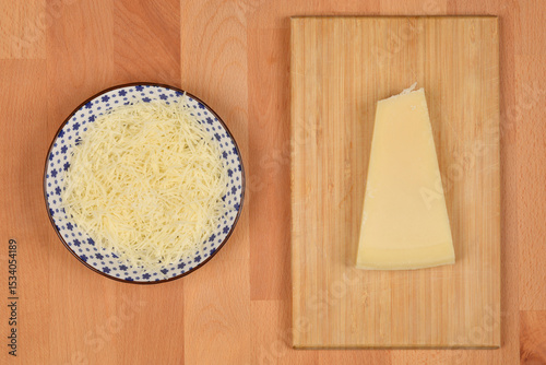 A top-down view displays a bowl of grated cheese beside a triangular wedge of cheese on a wooden cutting board, all on a wooden background
