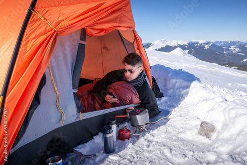 Man resting inside orange tent on snowy mountain, using smartphone. Camping stove, thermos, and cup on snow in foreground. Winter camping, mountain adventure, outdoor survival