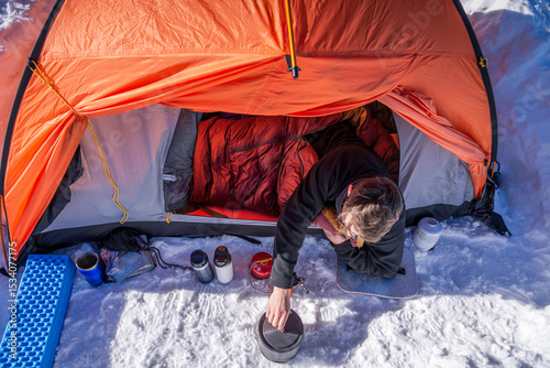 Man lying in sleeping bag cooks on portable stove outside winter tent in snowy campsite.Top-down view of cold weather camping gear