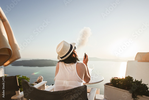 A woman enjoys a scenic sunset view over the Aegean Sea from a Santorini terrace with a glass of wine and a vape.