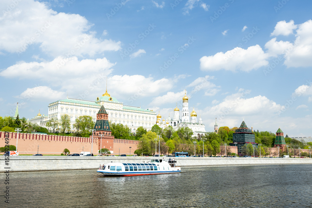 Fototapeta premium A scenic view of the Moscow Kremlin along the river. The historic architecture features towers and domes under a clear blue sky with fluffy clouds. A boat sails on the water.