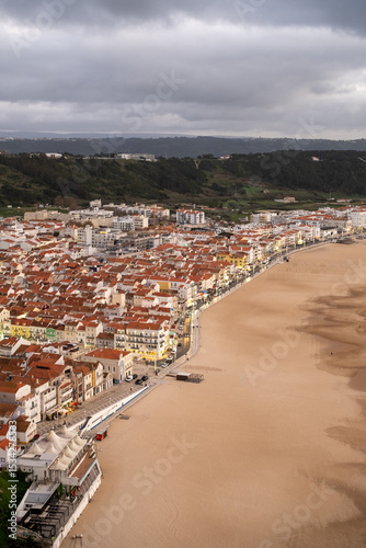 Aerial view of Nazaré town and beach in Portugal with red rooftops and evening lights under heavy clouds. The image shows the urban layout along the wide sandy coast.