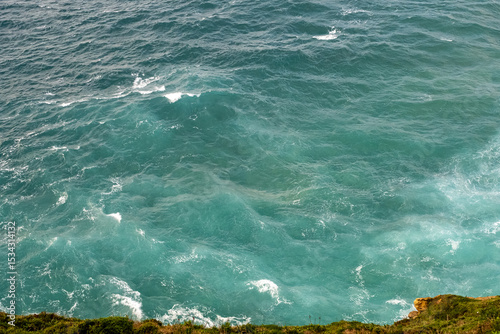 Three seagulls soar over textured turquoise waves of the Atlantic Ocean near a rocky coastline. Shot from above, the photo captures the rhythm and color of open sea and bird movement.