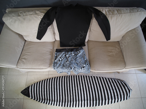 Black wetsuit and swim trunks lie on a couch next to surfboard, suggesting preparations for a surfing.