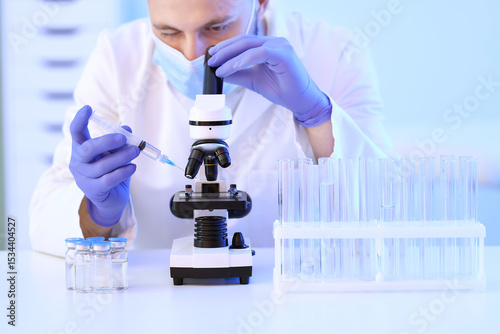 Male scientist with syringe and ampules looking through microscope in laborat...