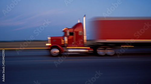 Red semi truck speeding down a highway at dusk with motion blur and a blue sky in the background