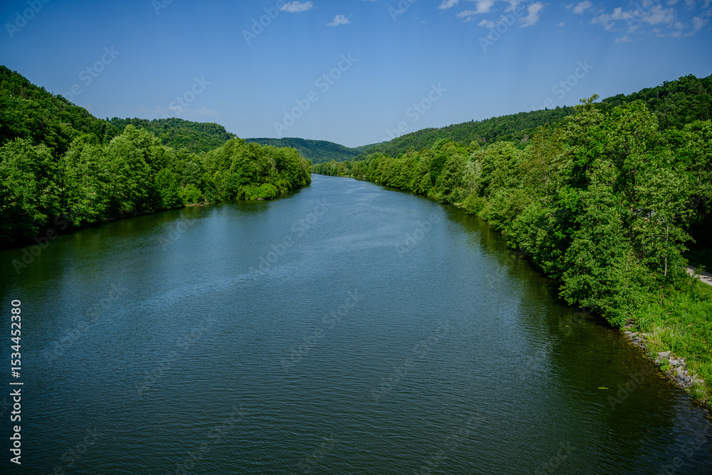 Fototapeta premium Serene river flows through lush green hills in a tranquil landscape with blue skies and fluffy clouds