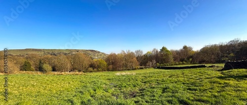 Fototapeta Naklejka Na Ścianę i Meble -  Expansive green meadow under a clear blue sky, framed by a distant hills. Sunlight deepens contrasts in the serene fields near Beechwood Park, Halifax, Yorkshire, UK