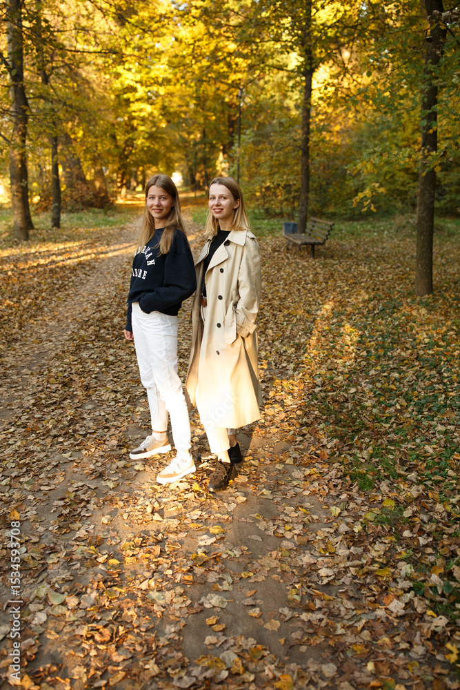 Fototapeta premium Two friends pose together in a golden autumn park, enjoying the warm light and cozy atmosphere of a beautiful day.