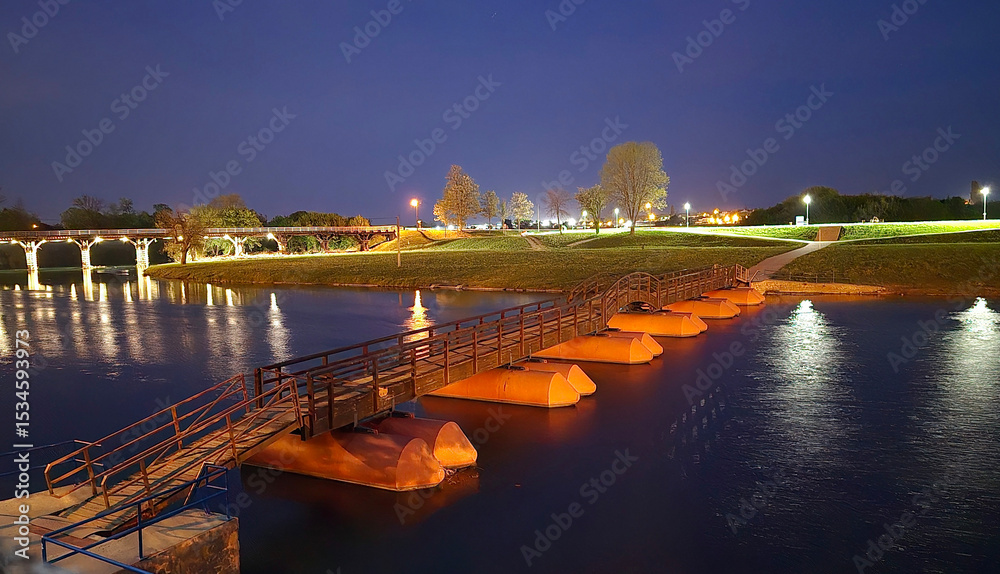 Fototapeta premium Floating pontoon footbridge with orange floats crosses a calm river, glowing under streetlights, with grassy banks and a historic arched bridge lit in the background on a clear night.