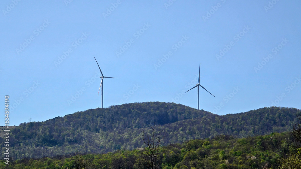 Two large wind turbines with three blades each stand atop a densely forested hill, generating renewable energy against a bright blue sky, surrounded by spring foliage and distant power lines. Two large wind turbines with three blades each stand atop a densely forested hill, generating renewable energy against a bright blue sky, surrounded by spring foliage and distant power lines.