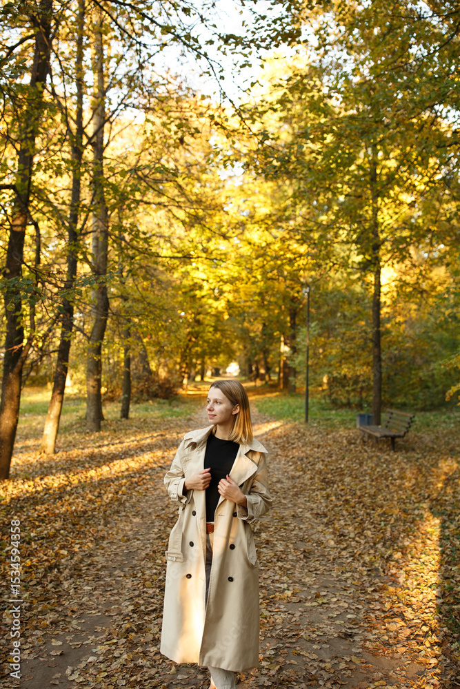 Fototapeta premium Stylish young woman in beige trench coat standing on a forest path during golden autumn sunset. Calm atmosphere, fashion look, casual outdoor lifestyle in nature.
