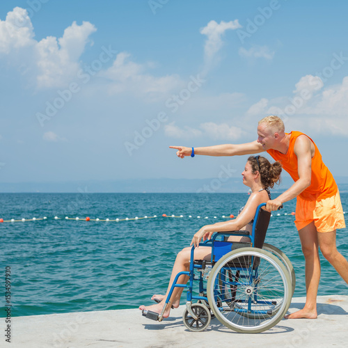 Smiling woman with a disability and her assistant helping her wheelchair move, enjoying time at the beach and the sea horizon view.